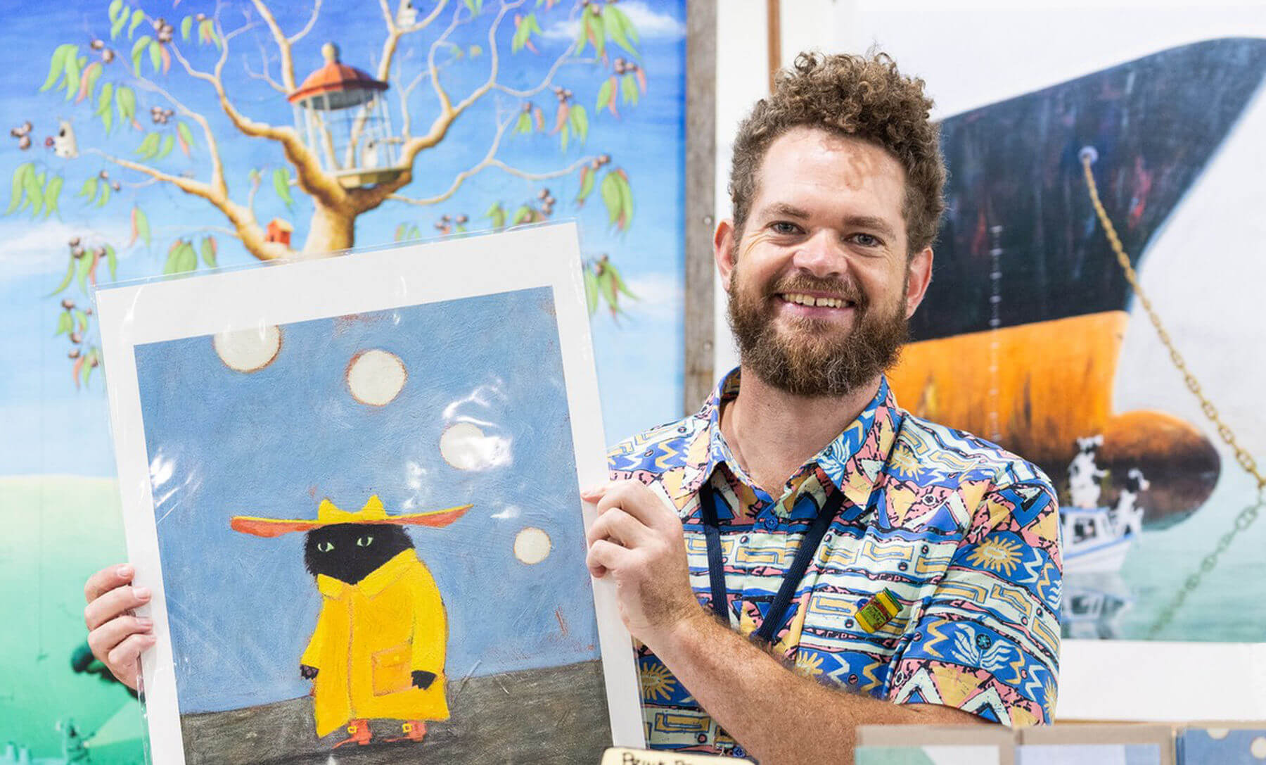 Australian Artist Beau Wylie holding a painting of a cat in a yellow raincoat with paintings in the  background
