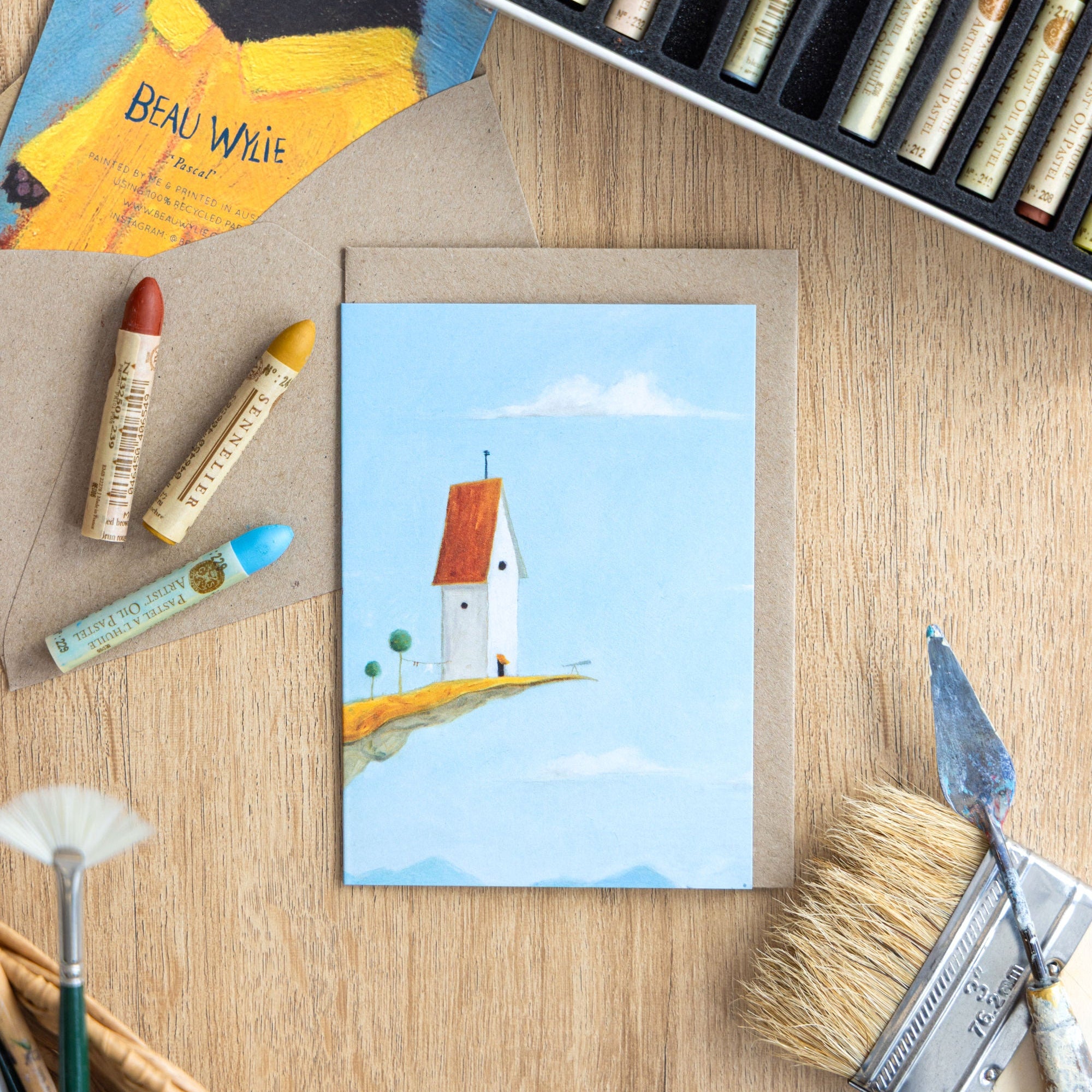 Art supplies including pastels, a paintbrush, and a greeting card with a lighthouse illustration on a wooden surface.